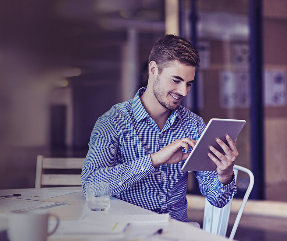 Foto de um homem sentado apoiado na mesa enquanto utiliza o tablet.