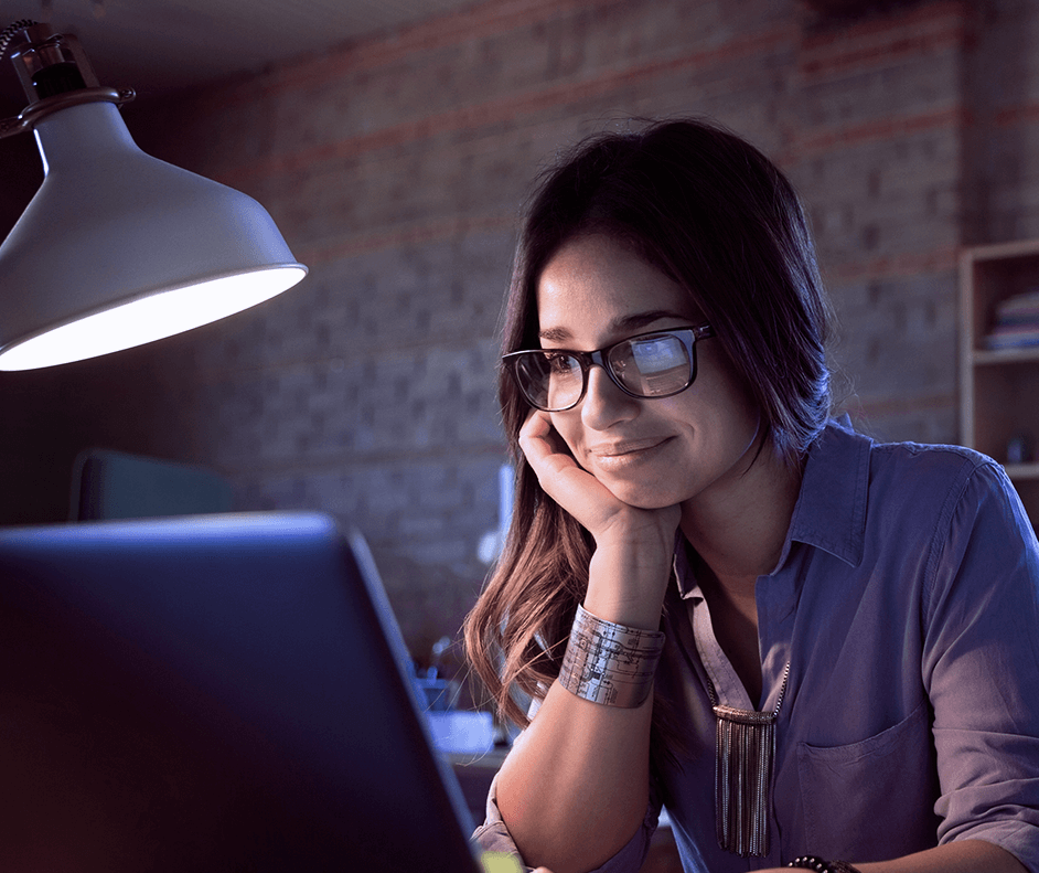 Fotografia de uma mulher mexendo em um notebook, numa sala iluminada apenas por um abajur.
