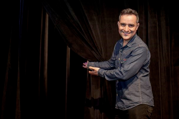 Foto do curador André Acioli em um palco com os movimentos de braço sugerindo um convite ao teatro.
