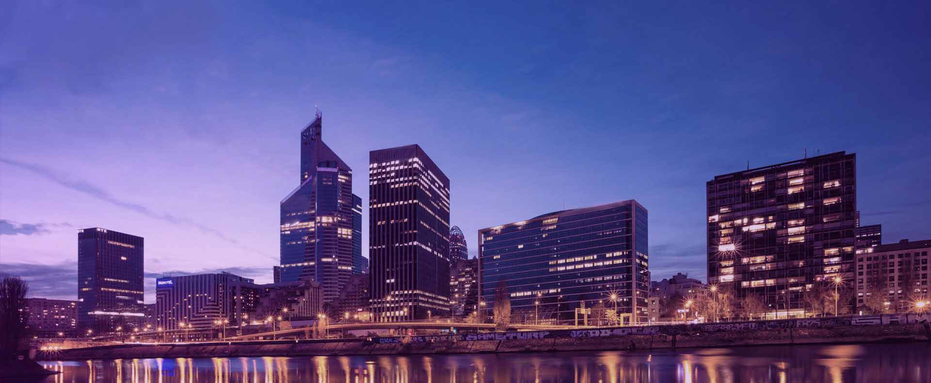 Imagem do skyline de uma cidade no início da noite, com diversos prédios acesos.