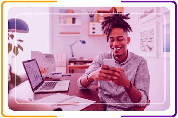 Imagem de um rapaz sorrindo sentado na mesa de trabalho e navegando no celular