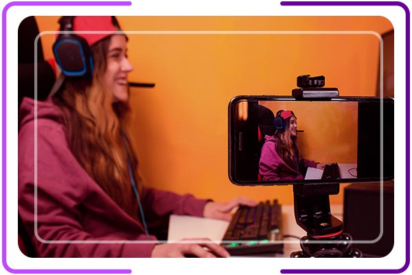 Imagem de uma jovem em um computador. Em primeiro plano, um celular a filmando.
