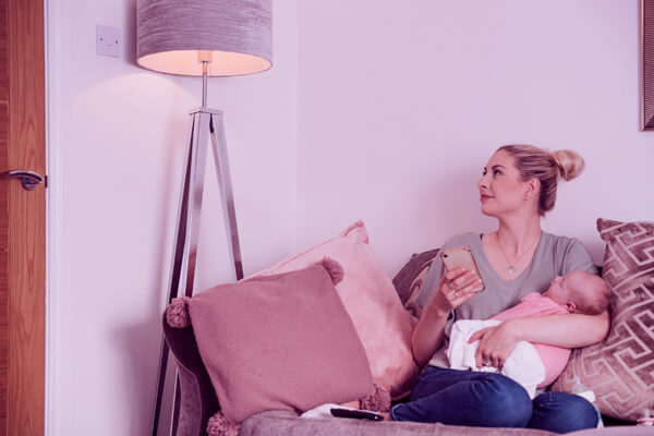 Imagem de uma mulher com um bebê no colo, ajustando a luz de um abajur via smartphone