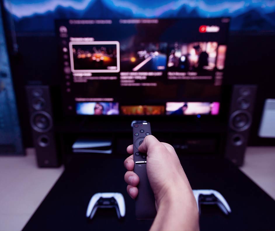 Pessoa apontando controle inteligente para Smart TV. Ao fundo, Xbox com dois controles de videogame