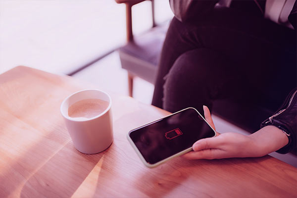 Mulher segurando celular com ícone de bateria acabando e um copo de café ao lado na mesa