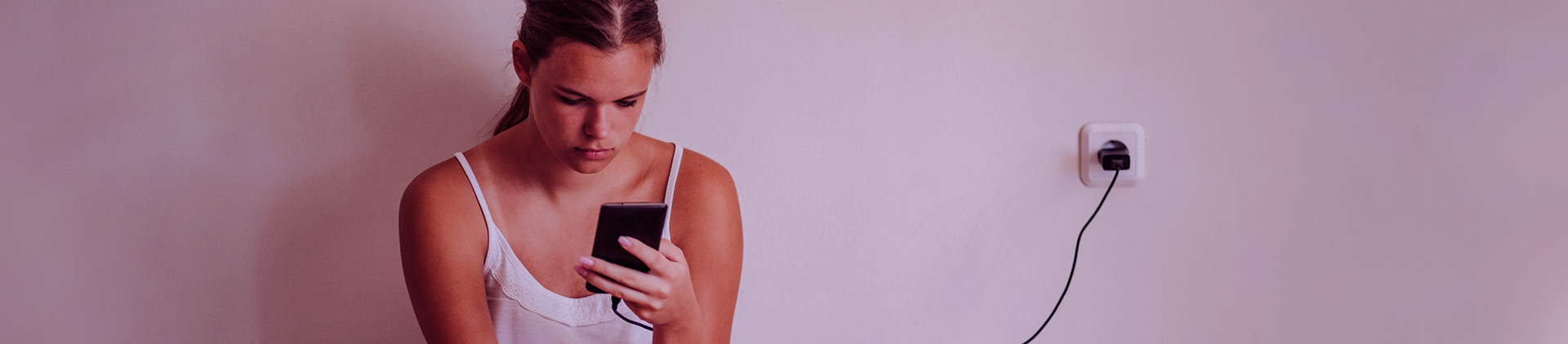 Garota jovem, com uma blusa branca, com o olhar frustrado para o celular na tomada que não carrega