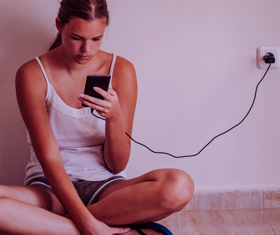 Garota jovem, com uma blusa branca, com o olhar frustrado para o celular na tomada que não carrega