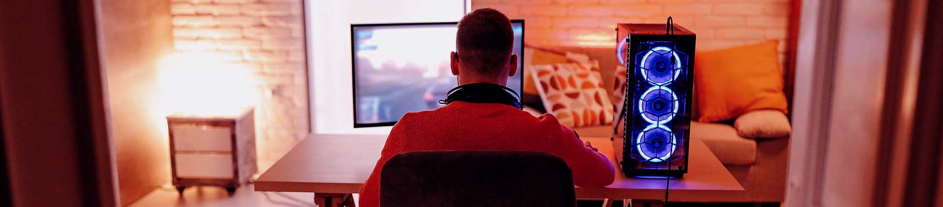 Pessoa, gamer, jogando no computador. Na imagem, mesa está ao centro com setup e, ao fundo, sofá.