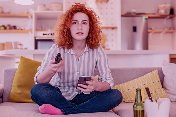 Jovem sentada no sofá de casa com o controle remoto numa mão e o celular em outro.