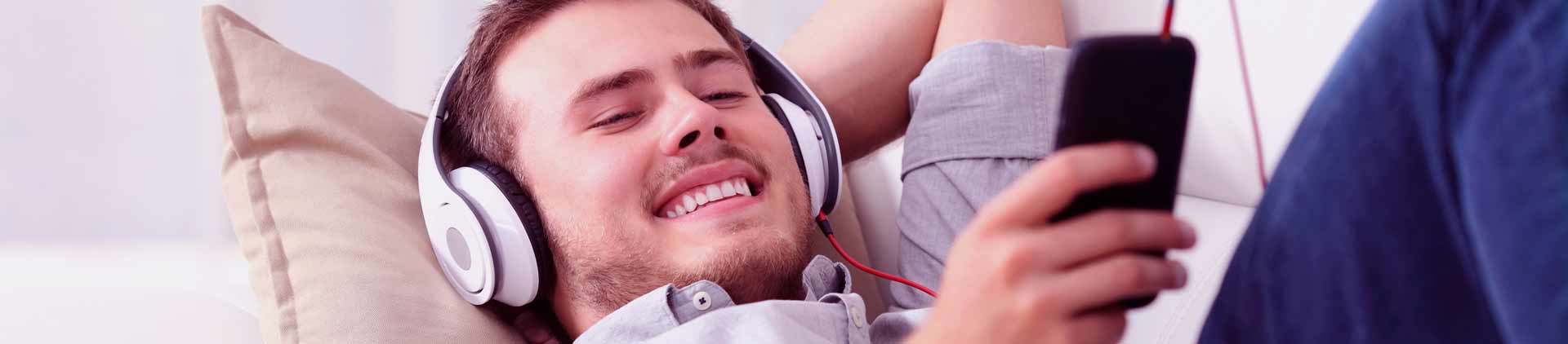 Homem deitado no sofá assistindo vídeo no celular com fone de ouvido. Parece feliz.