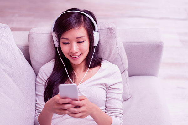 Mulher deitada no sofá assistindo vídeo no celular com fone de ouvido. Parece feliz.