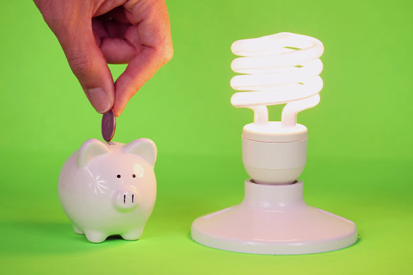 Mão colocando uma moeda dentro do cofre de porquinho e ao lado uma lâmpada LED