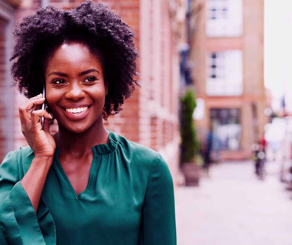 Mulher negra de blusa verde com o celular no rosto. Ela está ligando à cobrar e parece feliz