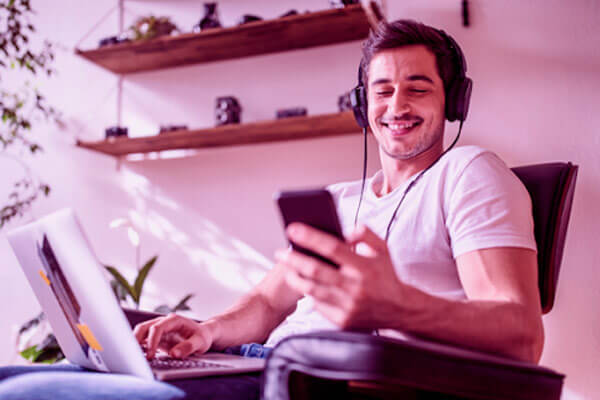 Homem sorrindo com headphone sentado em uma cadeira segurando um celular e com um notebook no colo.