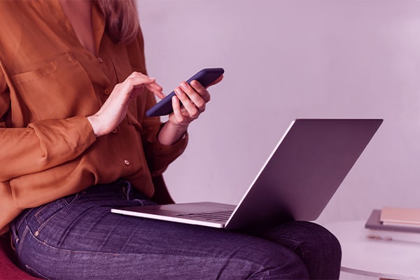 Imagem de uma mulher com um notebook no colo e o smartphone nas mãos.