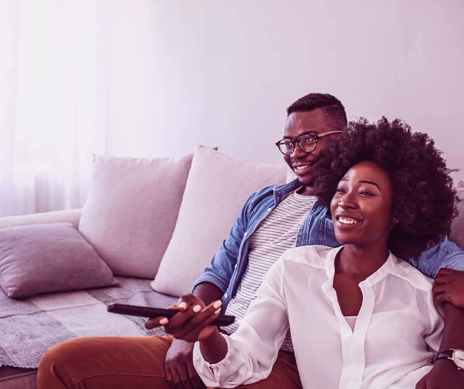 Casal abraçado no sofá. A mulher está segurando o controle da TV, procurando onde assistir doramas