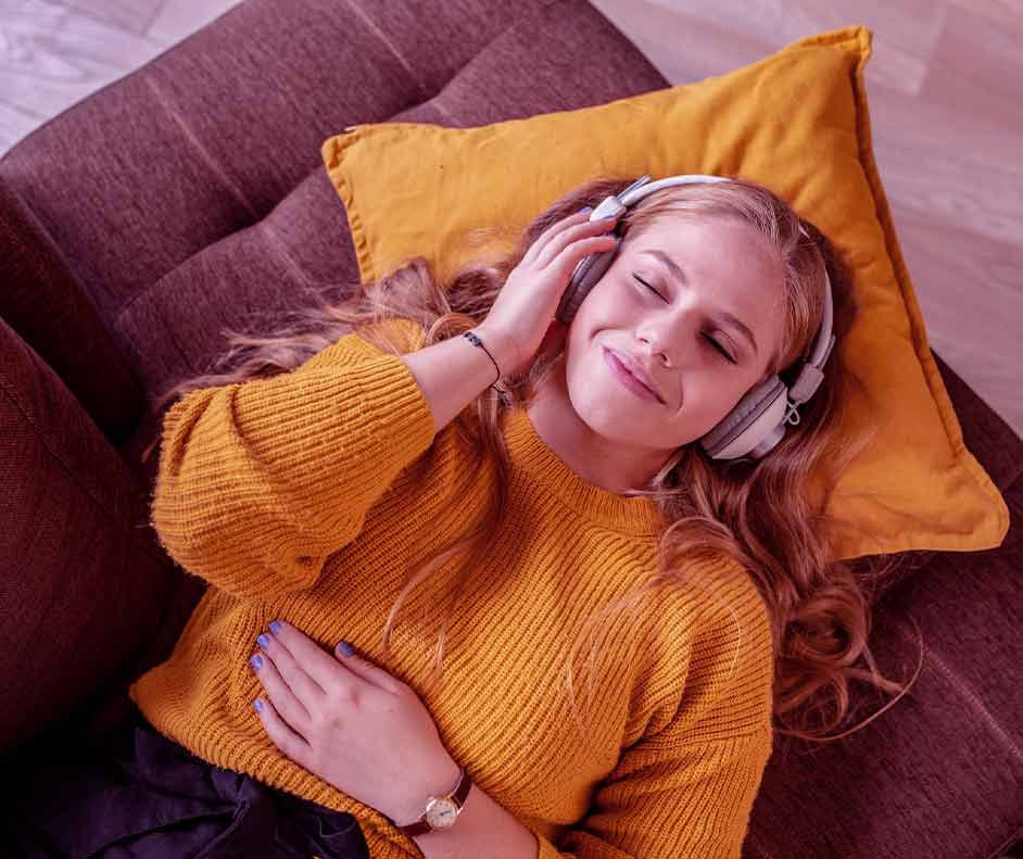 Mulher loira deitada no sofá de olhos fechados com headphone. Seu semblante mostra tranquilidade.