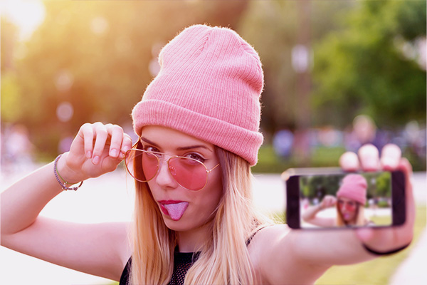 Imagem de uma jovem loira, com toca e óculos de sol rosas  fazendo careta tirando uma selfie. 