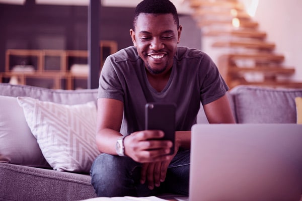Homem sentado no sofá e usando o celular e o notebook em seu apartamento