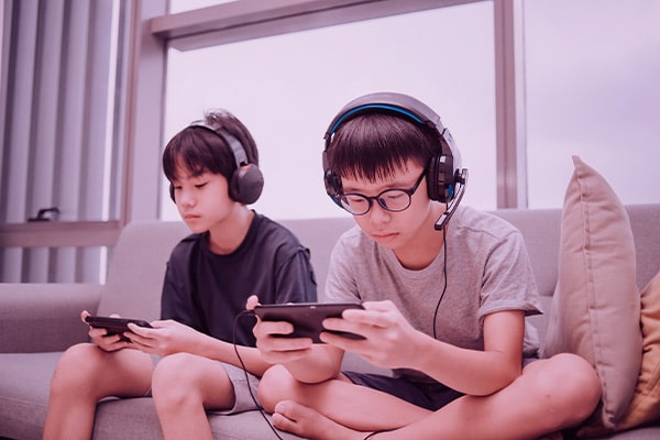 Duas crianças sentadas no sofá jogando no celular e usando fone de ouvido