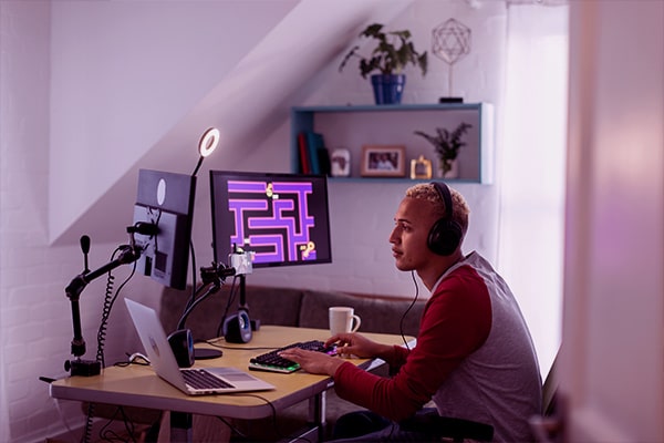 Jovem sentado na mesa e jogando com headset, notebook e duas telas a sua frente