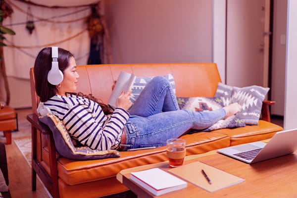 Moça deitada no sofá e lendo um livro enquanto escuta música para concentração no headphone