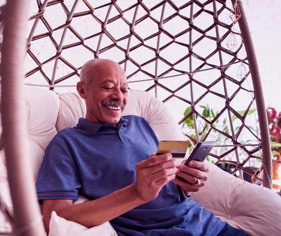 Homem de mais idade feliz, sentado no sofá, segurando o cartão e fazendo uma compra no celular.
