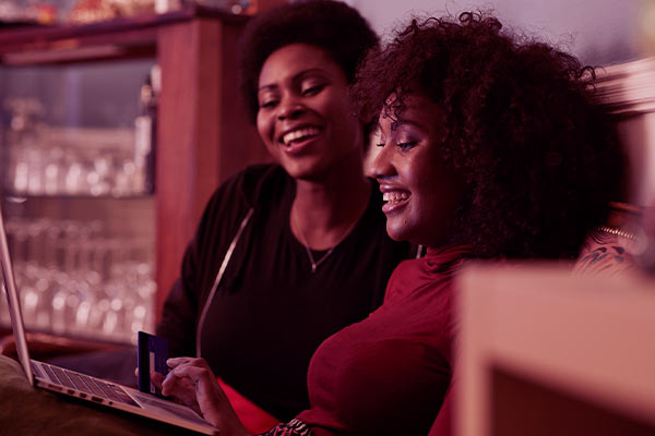 Duas mulheres comprando em um notebook enquanto uma delas segurando um cartão de crédito
