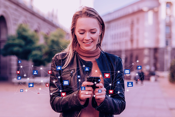 Imagem de uma mulher usando o celular na rua. Sobre o telefone, flutuam ícones de redes sociais.