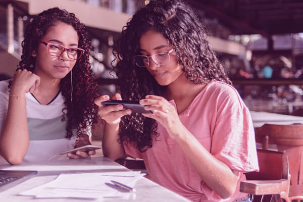 Imagem de duas mulheres sentadas à mesa, onde há um notebook. Uma delas olha para a tela do celular.