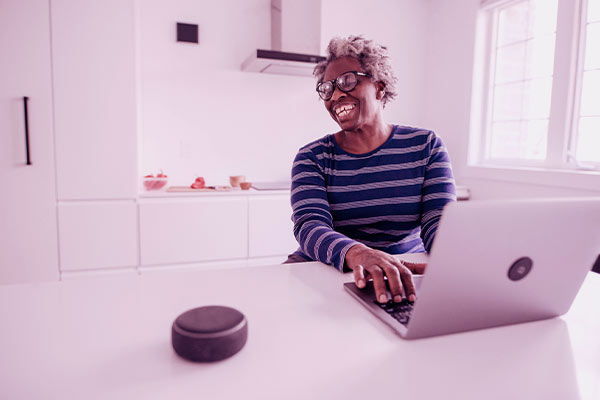 Senhora sorridente ao se comunicar com a Alexa em sua cozinha e com o notebook. 