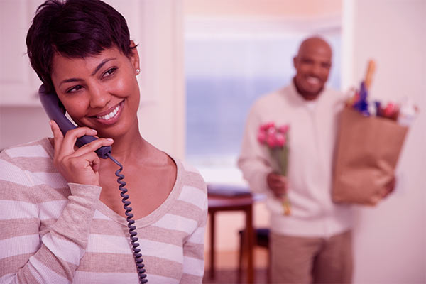 Mulher conversando no telefone fixo enquanto um homem chega com as compras e flores atrás dela.