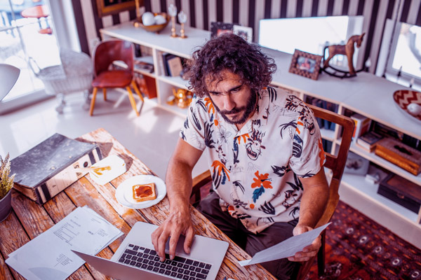 Imagem de um homem trabalhando no notebook. Na mesa, há um prato com uma torrada.