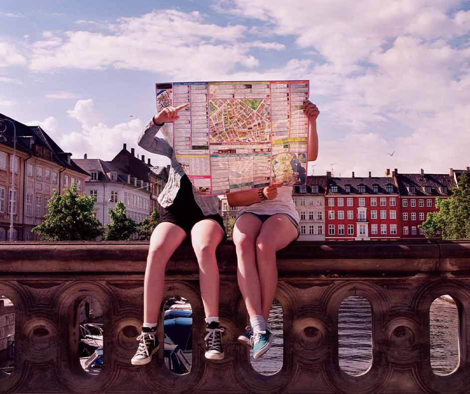 Duas mulheres sentadas em um muro baixo procurando locais para viajar sem gastar muito.