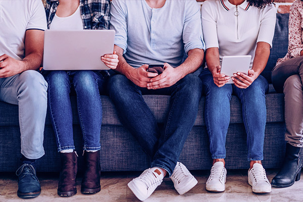 Imagem de um grupo de amigos sentados no sofá, dois deles seguram um notebook e um tablet.