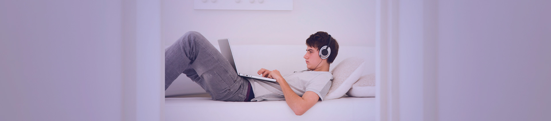 Imagem de um jovem sentado em um sofá, utilizando um notebook e headphones.